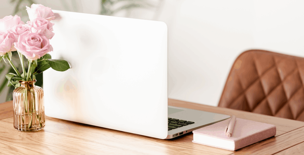 Photo of a desk with a laptop, flower vase, pen and notebook.