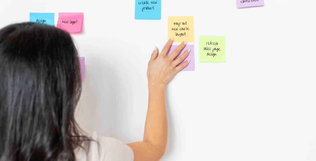 Alt text:
Overwhelmed entrepreneur organizing colorful sticky notes on a white wall, reflecting task overload and competing priorities in an online business.