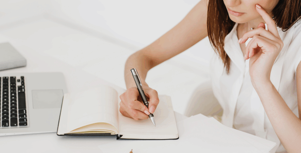 Woman planning in a notebook beside a laptop while deciding how to create systems before you're ready, representing entrepreneurs who are waiting to build systems because they feel unsure where to start or stop waiting to get organized.