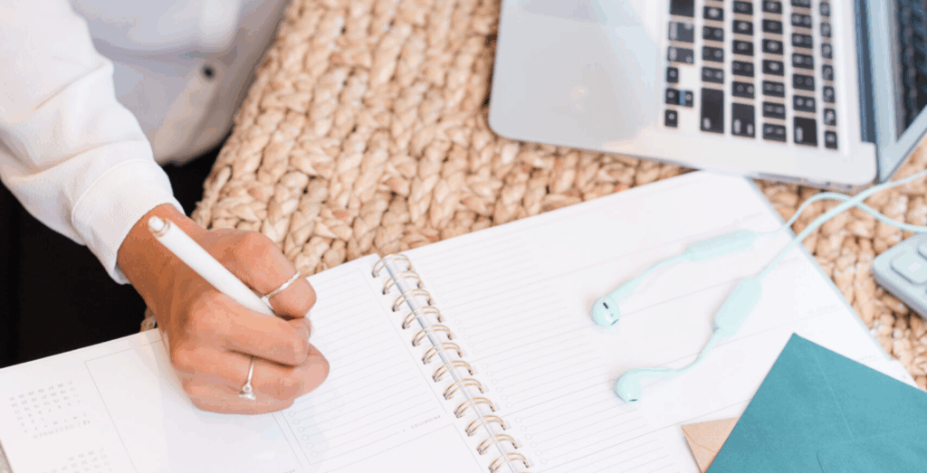 Entrepreneur writing in a planner beside a laptop and workspace setup, representing why you don't have systems in place and the challenge of prioritizing business systems while building systems for productivity and organization.
