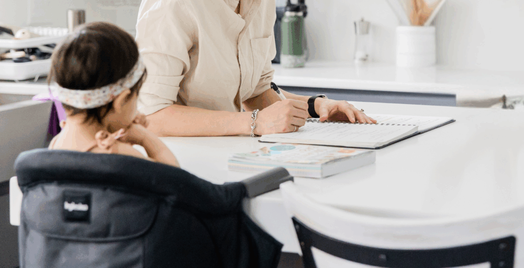 Solopreneur planning in a notebook while caring for a child at the kitchen table, representing feeling overwhelmed, creating simple systems, and managing everyday life as an overwhelmed solopreneur.